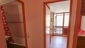 A hallway leading to a balcony with a wooden floor and a red and white patterned shower curtain.