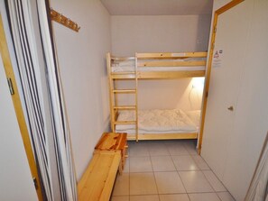 Une petite piÚce étroite, avec un lit superposé, un banc en bois et une porte.