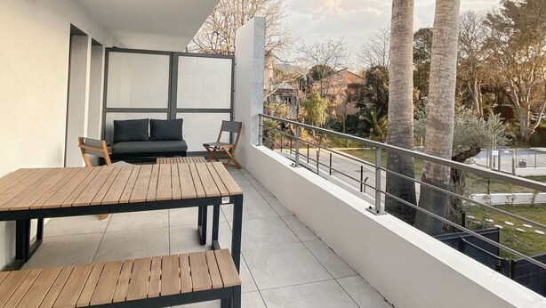 Un balcón con mesa y sillas de madera, un sofá, y vistas a palmeras y un jardín.