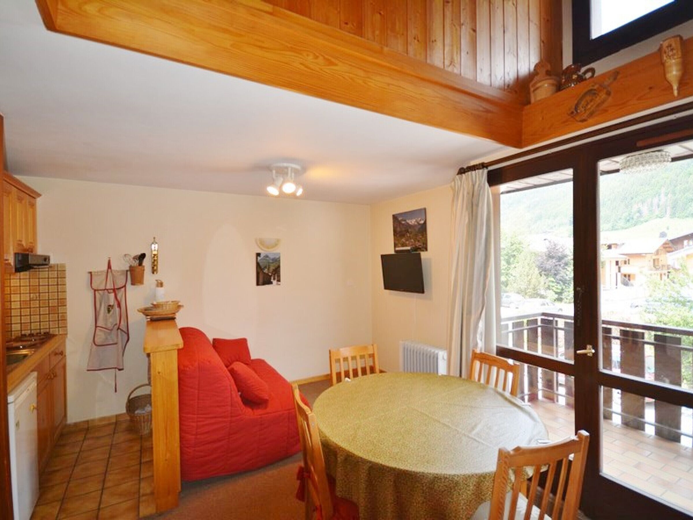 Um quarto aconchegante com teto de madeira, uma mesa de jantar com cadeiras, um pufe vermelho, uma área de cozinha com painel de azulejos e uma grande janela com vista para o exterior.