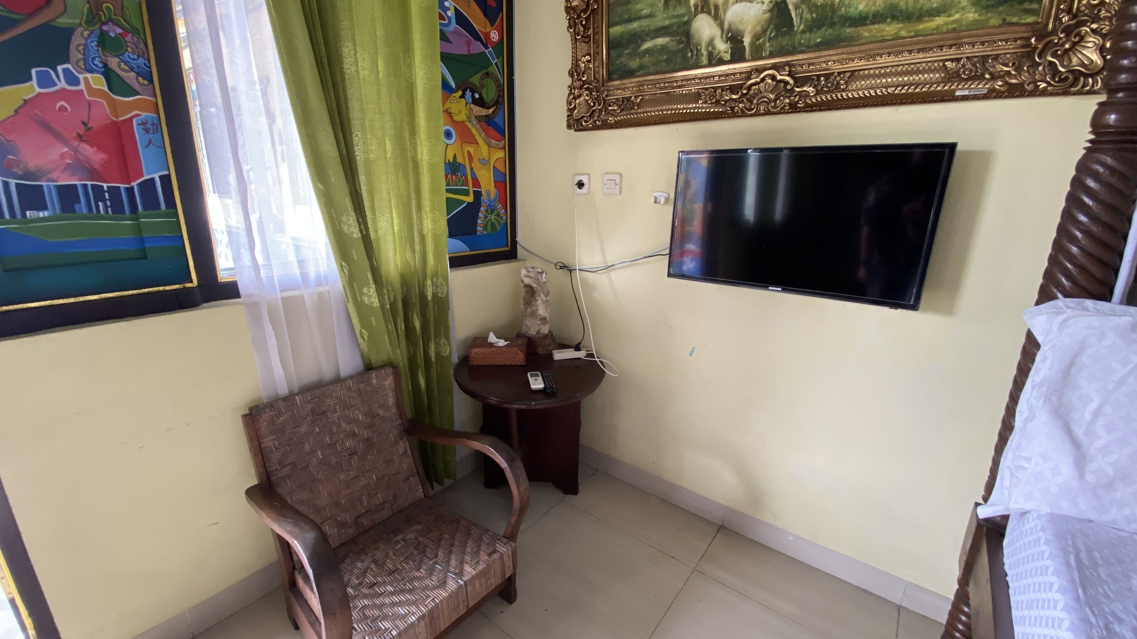 A room with a television mounted on the wall, a chair, a small table with a remote control, and a painting above the television.