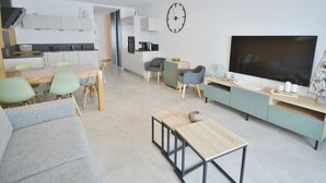 Een moderne woonkamer met een bank, een salontafel en een televisie. Er is een eethoek met een tafel en stoelen. Achtergrond is een keuken.