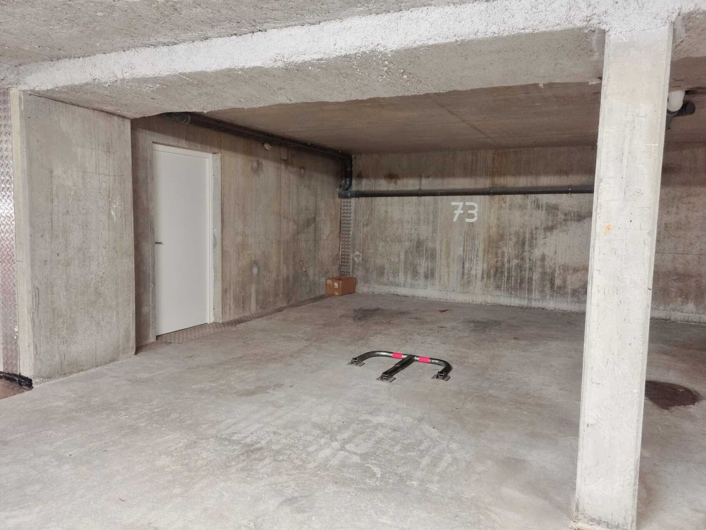 A concrete room with a door, a number '73' on the wall, and a black and pink object on the floor.