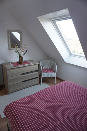 Um quarto com teto inclinado, cômoda com gavetas, cadeira com almofada, cama com colcha listrada e um claraboia.