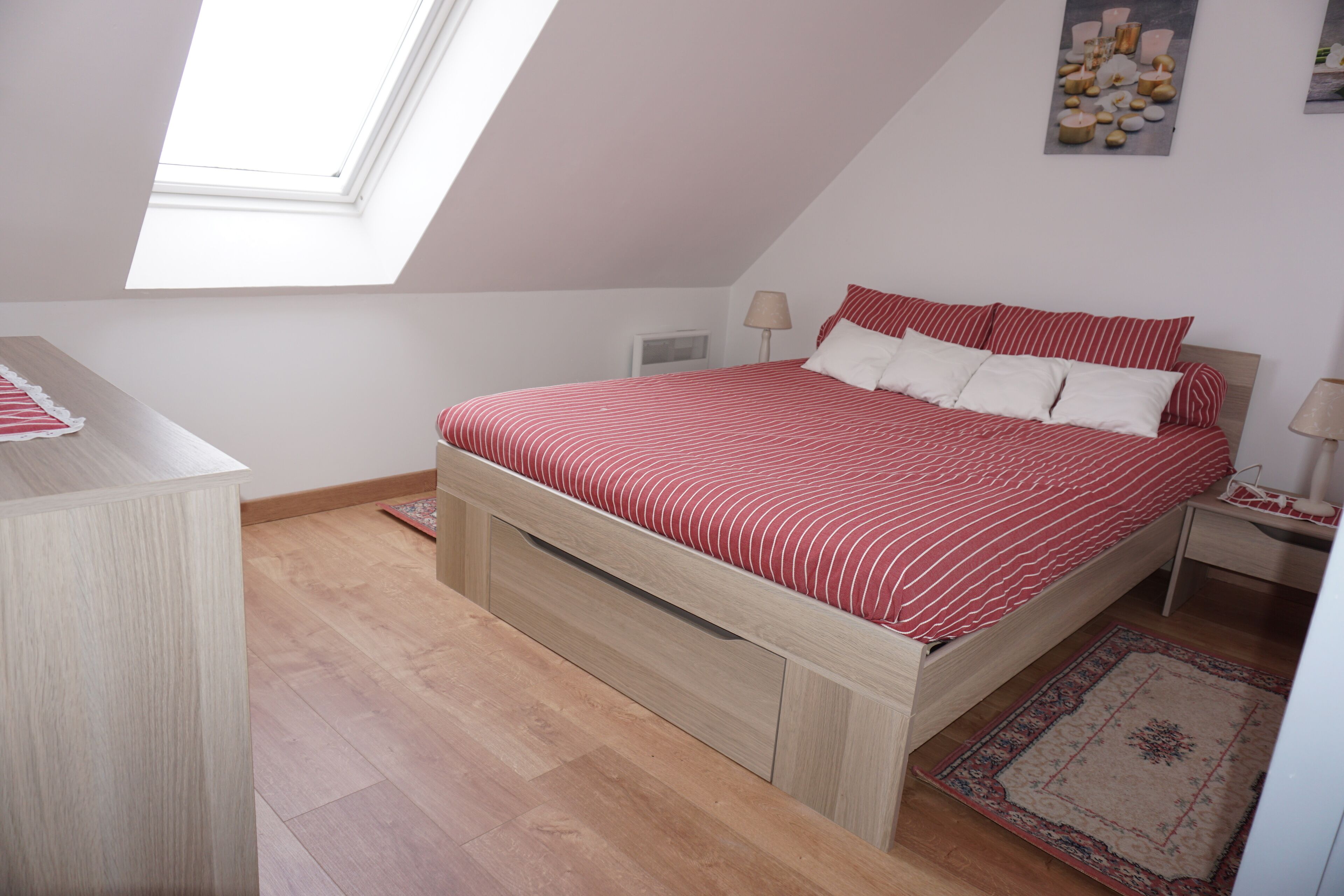 A bedroom with a sloped ceiling, a bed with red and white bedding, a wooden nightstand, a lamp, and a small rug.