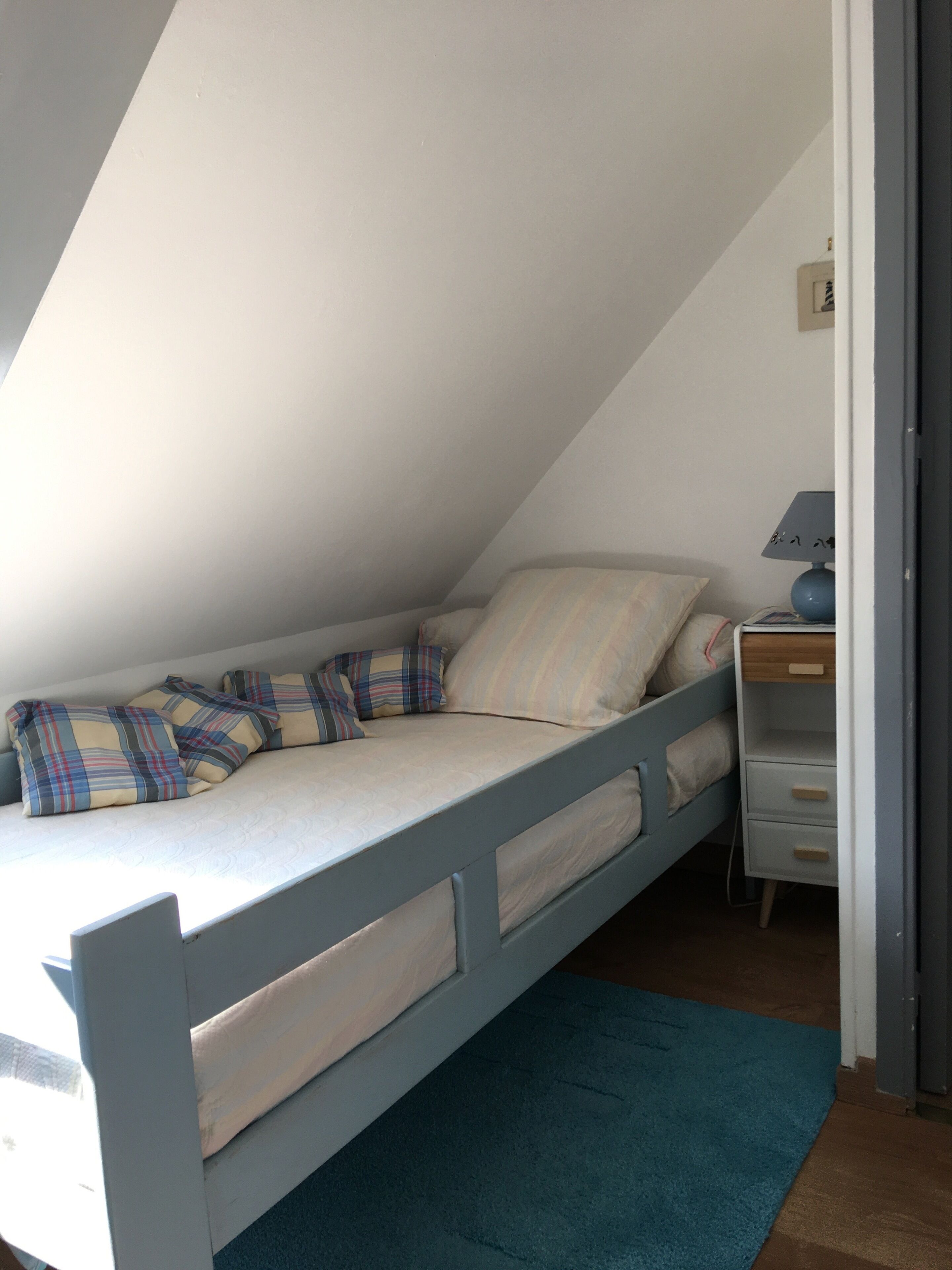 A small, single-bed room with a sloped ceiling, a bedside table with a lamp, and a blue rug.