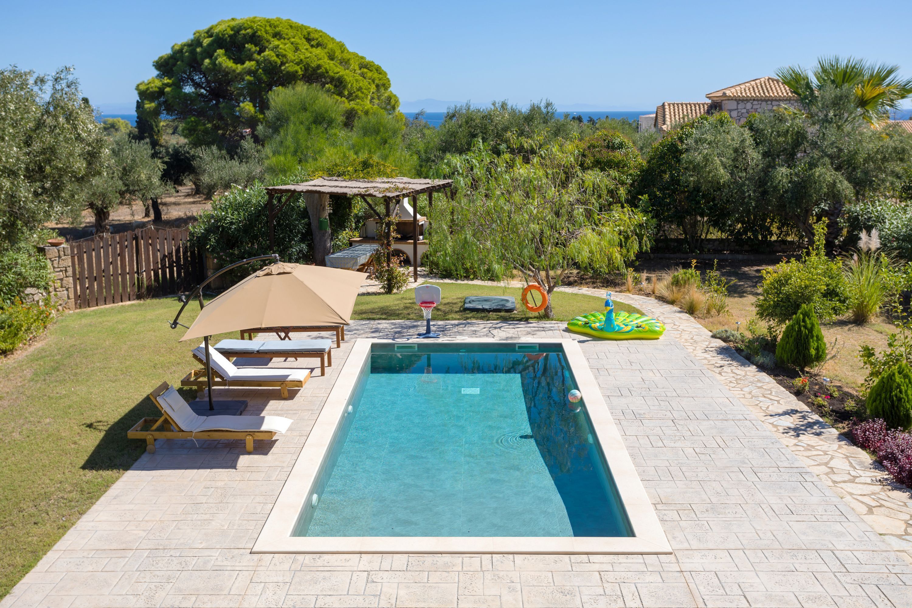 Una piscina rectangular con plataforma de baldosas, rodeada por una cerca de madera y una pérgola con toldo.