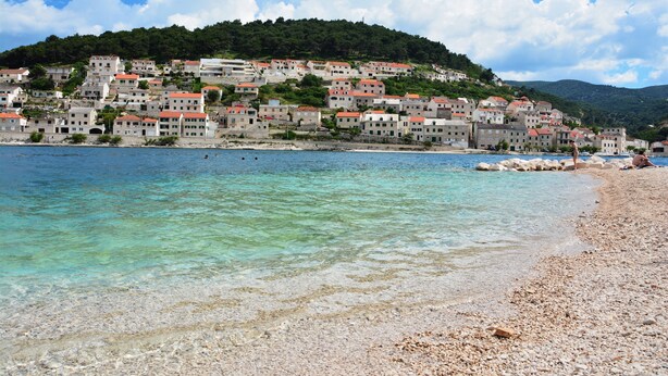 Eine Küstenstadt mit Häusern an einem Hügel, klarem türkisblauem Wasser und einem Kiesstrand.