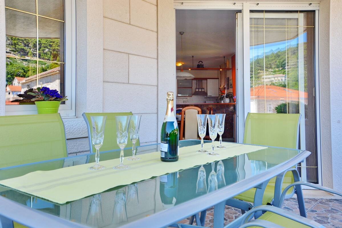 A glass table with a bottle and glasses, green chairs, and a view of a building through a window.