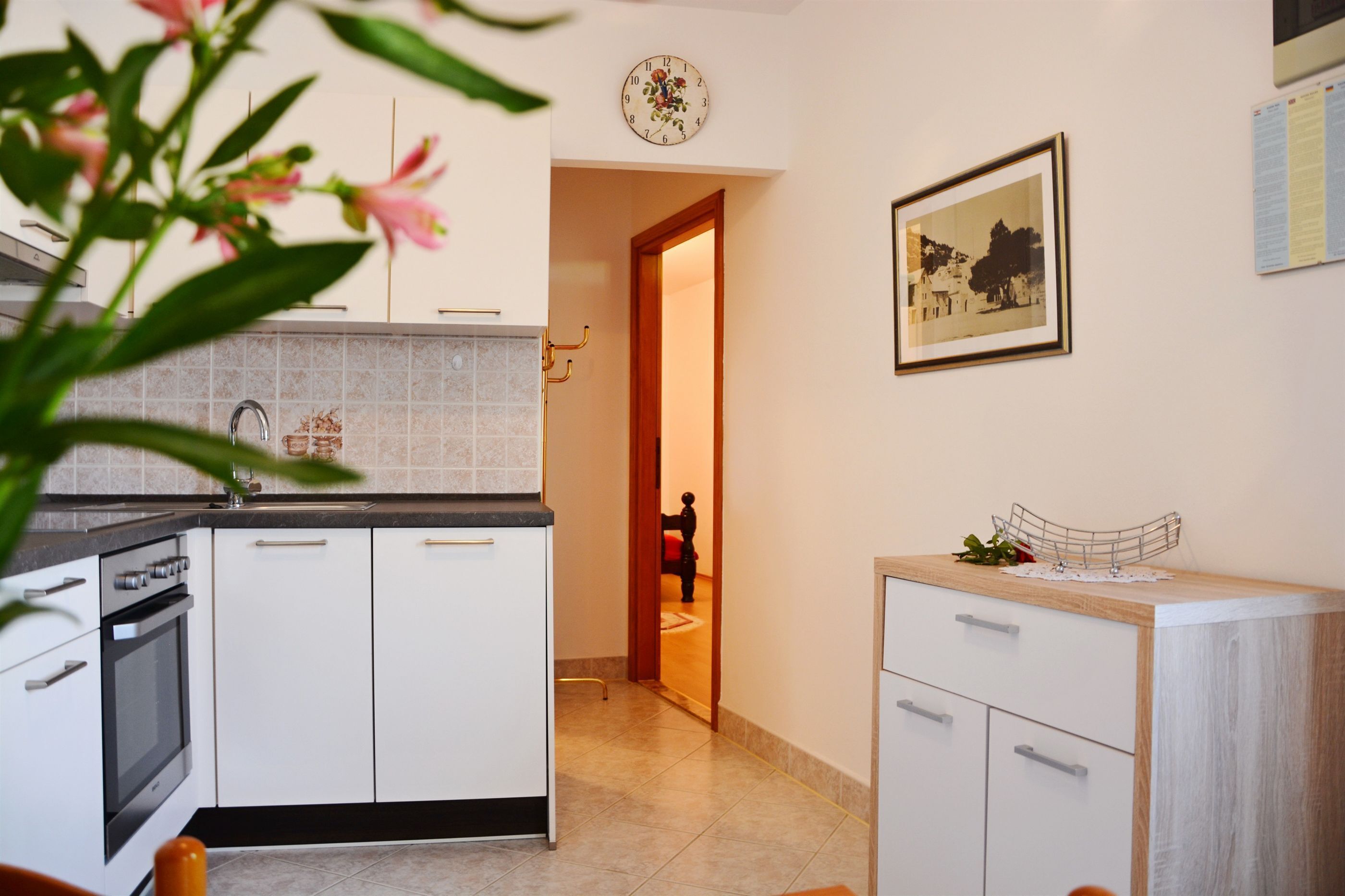 Eine moderne Küche mit weißen Schränken, einem Edelstahlbackofen und einer gefliesten Spritzschutzwand. Eine Holztür führt in einen anderen Raum, und ein gerahmtes Bild ist an der Wand angebracht.