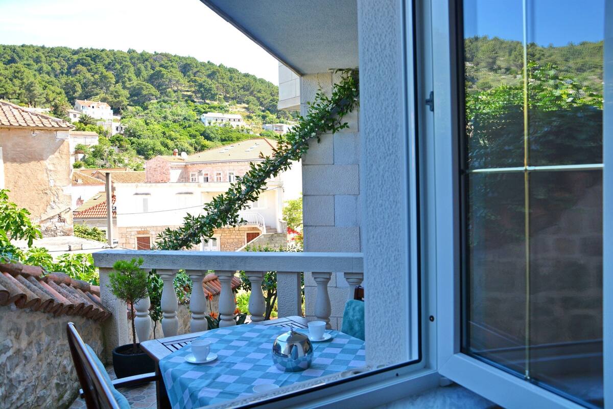 A balcony with a checkered tablecloth, a small potted plant, and a view of a hillside with buildings.