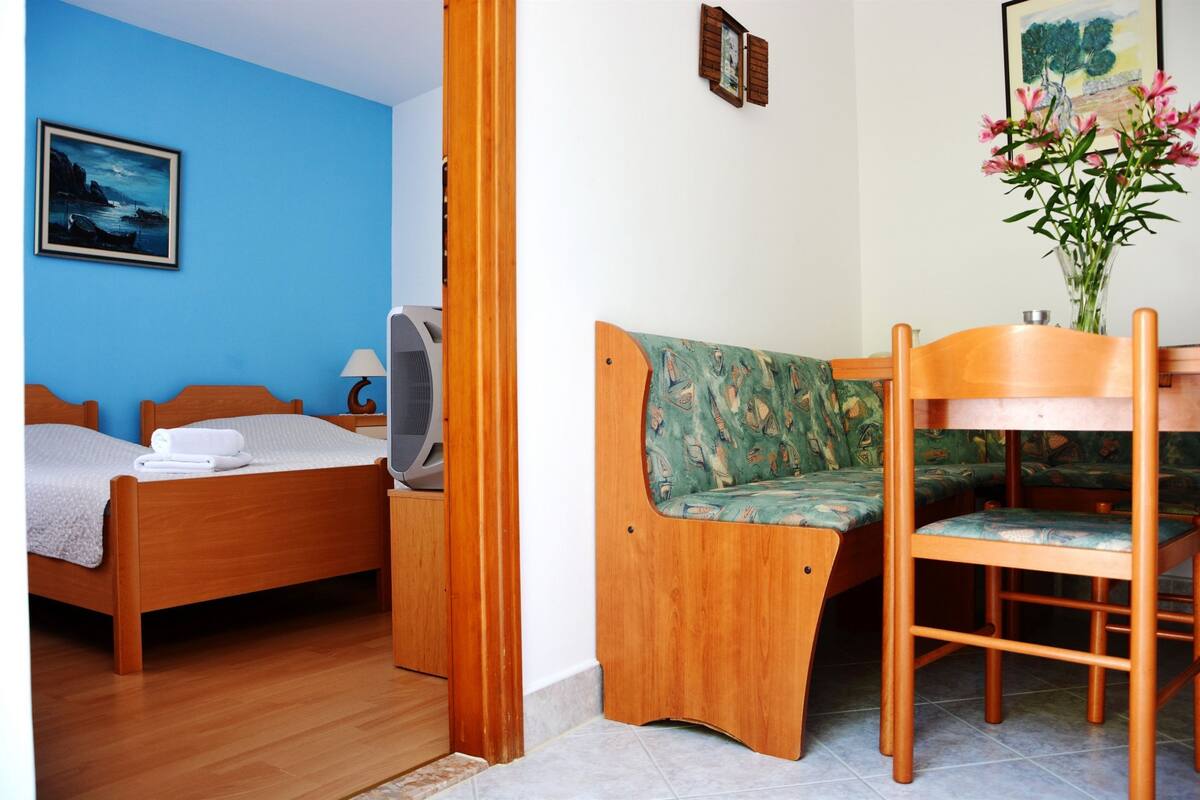 A room with a bed, a wooden chair, a table, and a vase with flowers.