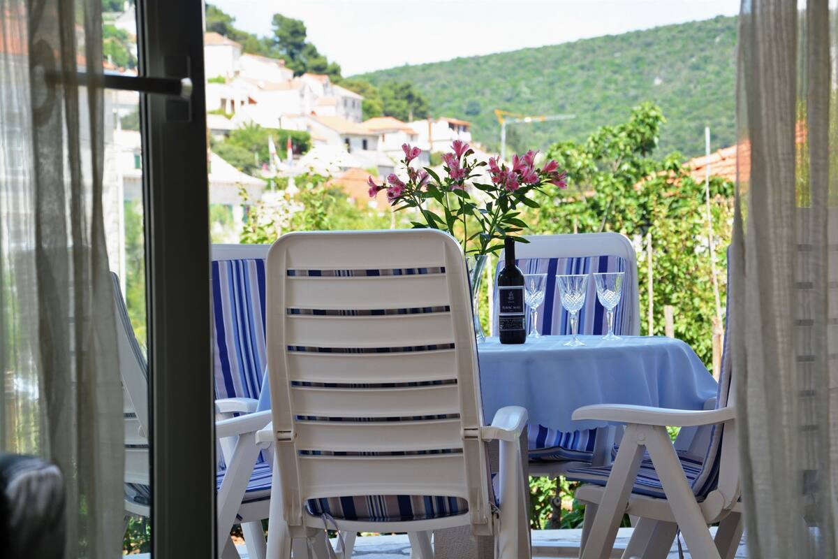 A balcony with two white chairs and a table, a vase with pink flowers, and a view of houses and greenery.
