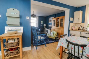 A room with a blue wall, wooden flooring, a dining table with chairs, a kitchen area with cabinets, and a hanging light fixture.