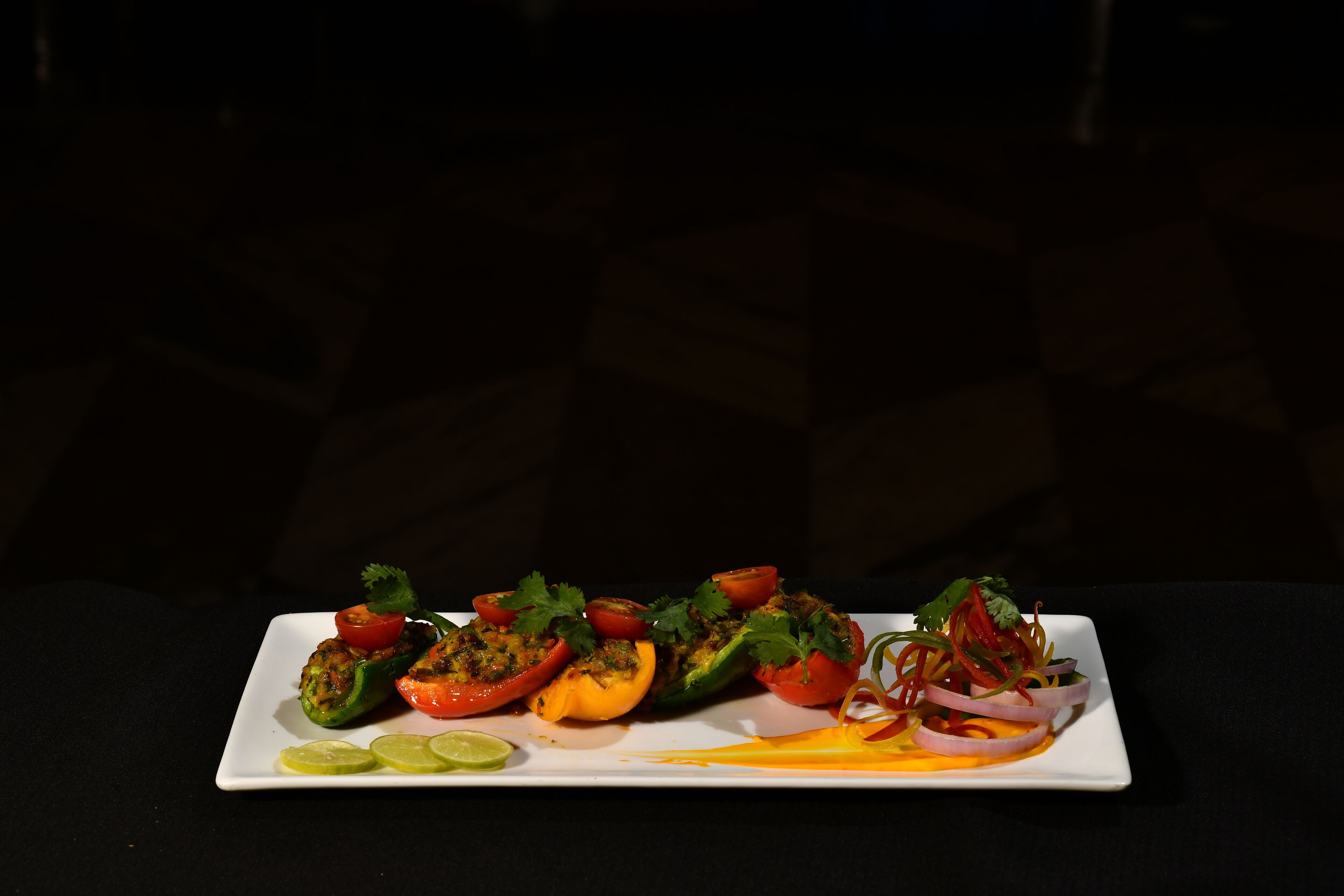 A square white plate with stuffed bell peppers, tomatoes, and a side of sliced onions and lime wedges.