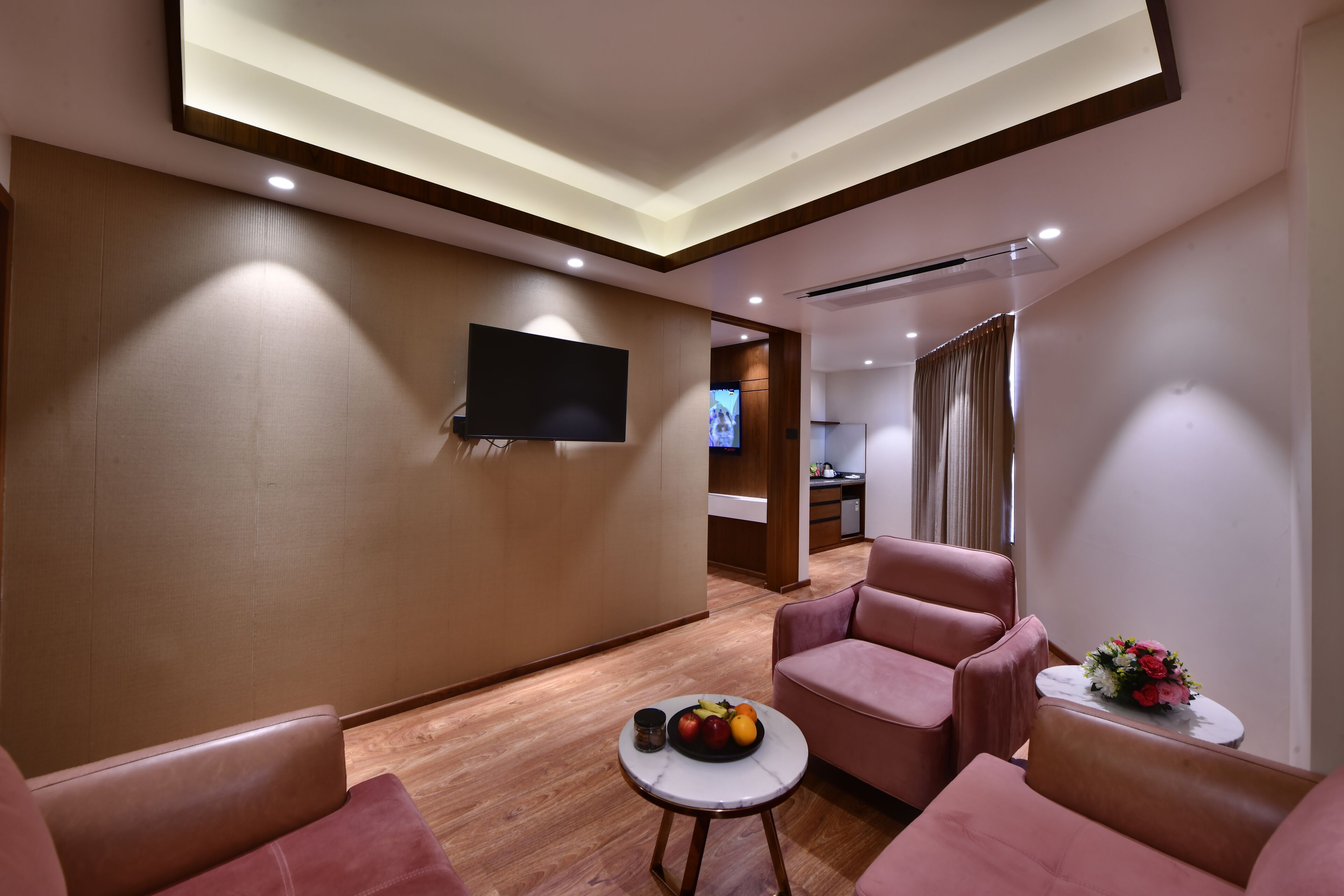A modern hotel room with a flat-screen TV mounted on the wall, a small round table with fruit, and two pink armchairs.