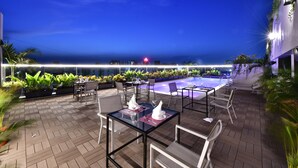 Un área de comedor al aire libre con mesas y sillas, una piscina y vistas al océano de noche.