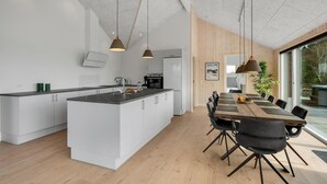 Et moderne køkken med en central ø, et spiseområde med et træbord og sorte stole, samt et stort vindue med udsigt til det fri.