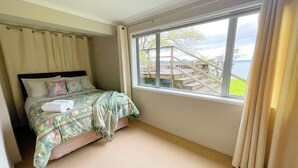 A bedroom with a bed, a window with a view of a deck and a body of water, and a curtain. - Lakeside Paradise (Rotorua)
