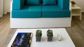 A modern living room with a teal sofa, a woven wall hanging, a small side table with a plant, and a book titled 'Tito Paris' on a white coffee table.