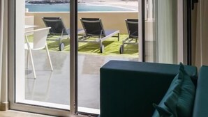 Ein Zimmer mit einer Schiebeglastür, die auf einen Balkon mit Blick auf Strand und Berge führt. Auf dem Balkon befinden sich eine grüne Couch und eine Lampe.