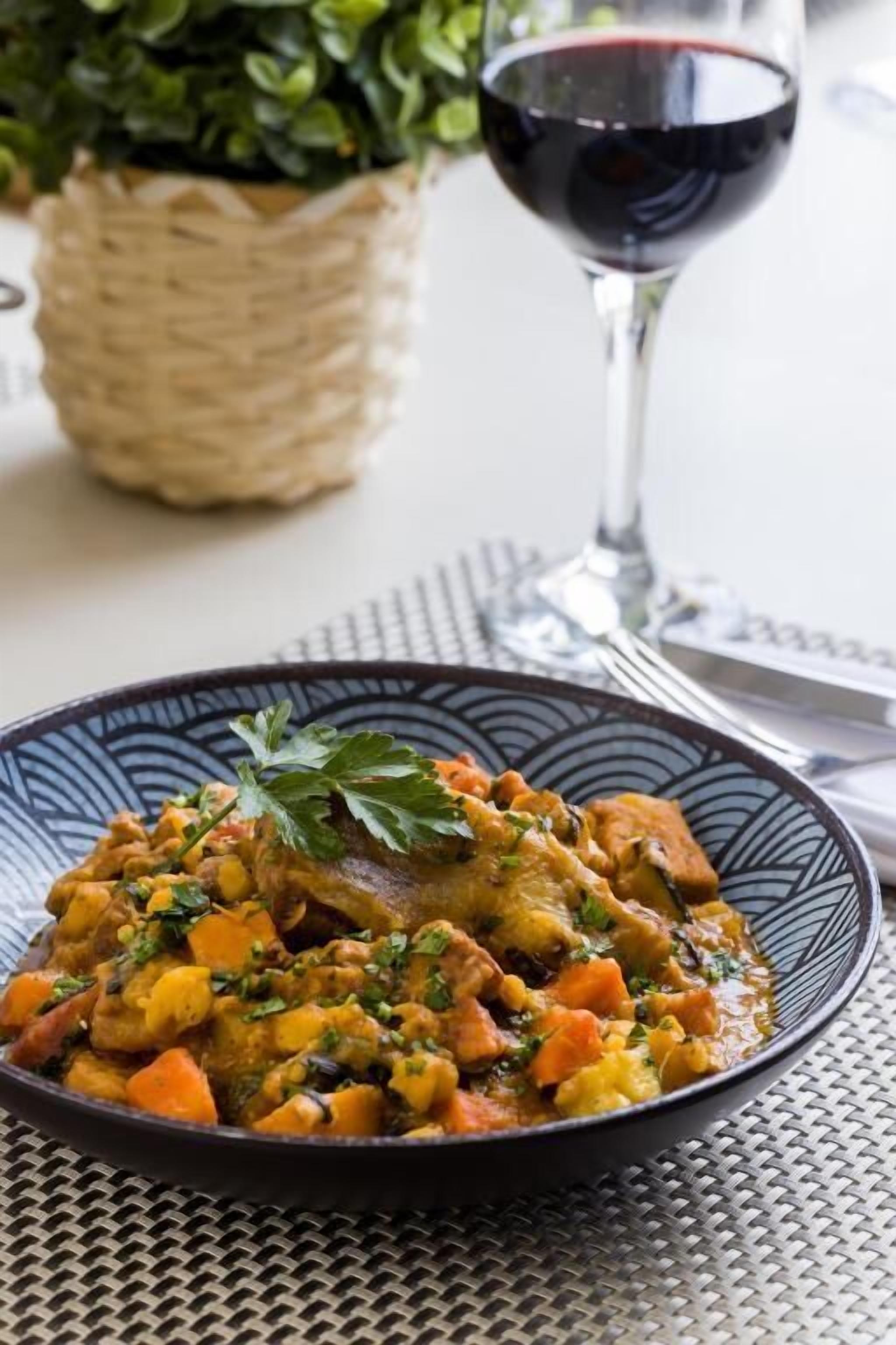 A plate of stew with vegetables and a glass of red wine on a table.