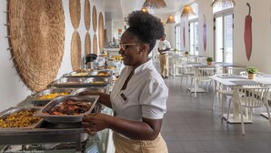 Ein Buffet mit verschiedenen Speisen, eine Person bedient die Gäste, und ein helles, modernes Interieur mit weißen Möbeln und Wanddekorationen.