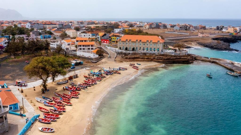 An aerial view of a coastal town with a beach lined with boats, buildings with orange roofs, and a clear view of the sea.