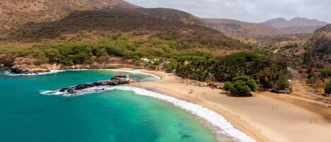 A coastal landscape with a sandy beach, clear turquoise waters, and a mountainous backdrop.