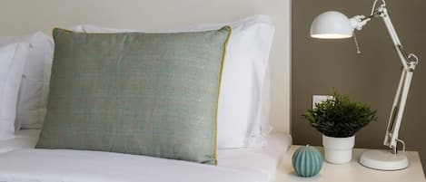 Una cama bien hecha con ropa blanca y una almohada color turquesa, una mesita de noche blanca con una lámpara y una planta en maceta, además de un pequeño globo decorativo.