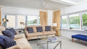 A spacious living room with a beige sofa set, a glass coffee table, and a view of a harbor through large windows.