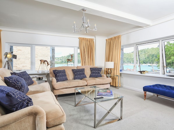 A spacious living room with a beige sofa set, a glass coffee table, and a view of a harbor through large windows.