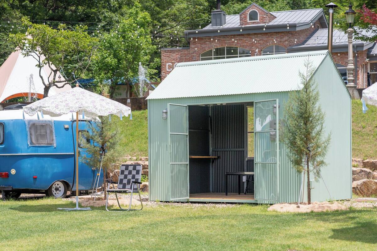 A blue vintage bus parked in a grassy area with a small shed and a house in the background.