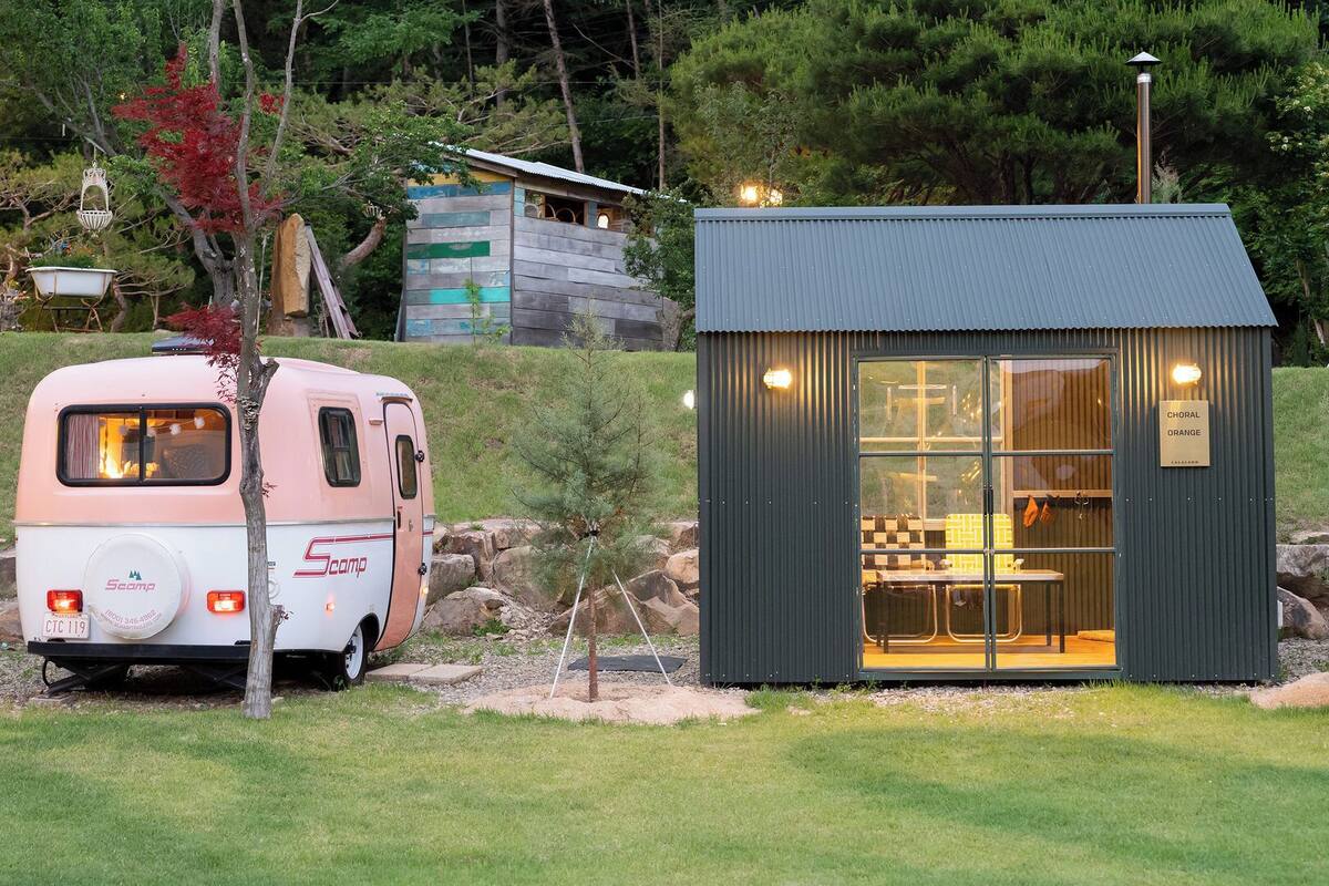 A Scamp RV parked beside a small, dark-colored building with a glass door and a sign reading 'CHORAL ORANGE.'