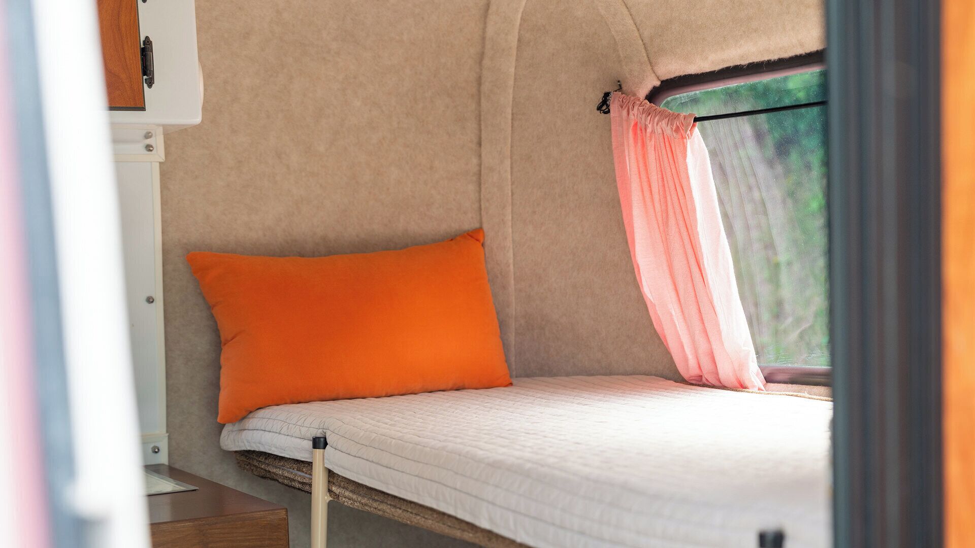 A single bed with a white mattress and an orange pillow in a tent-like structure with a window.
