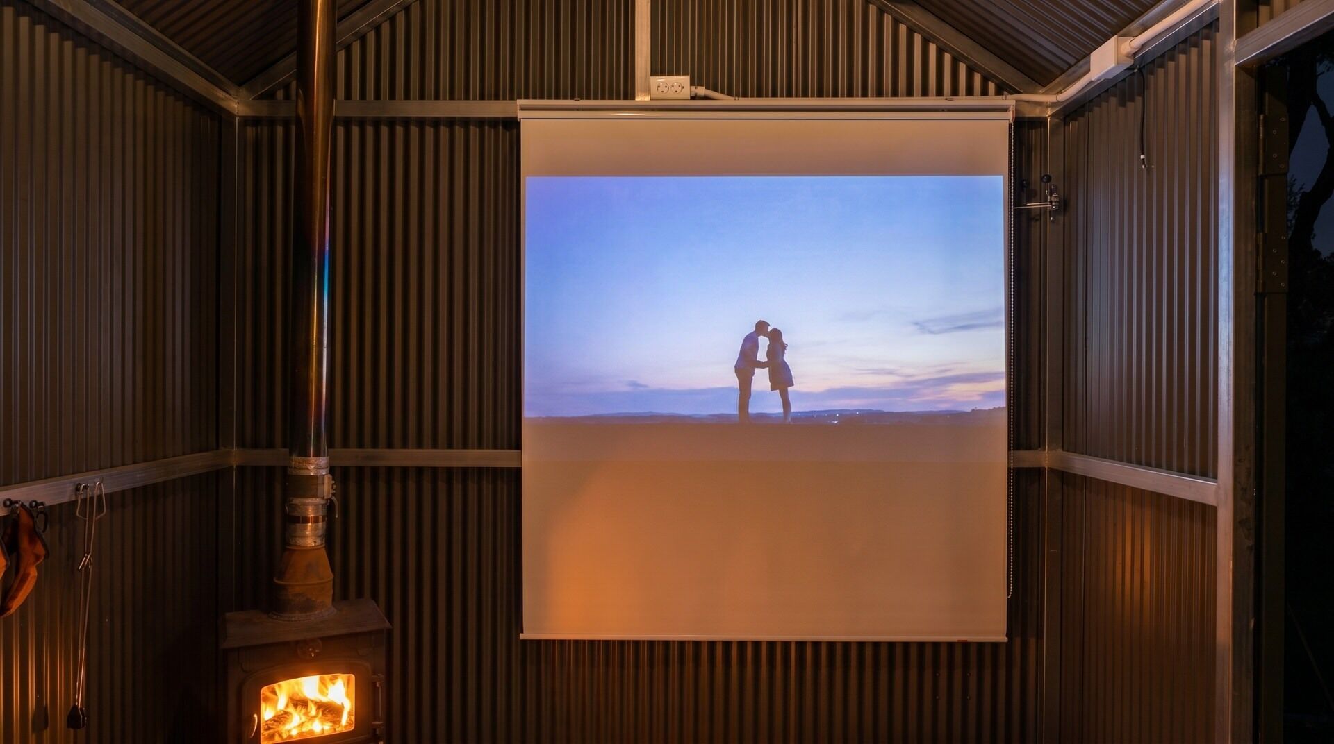 Una habitación con chimenea, pantalla de proyección y vista a una pareja en la playa.
