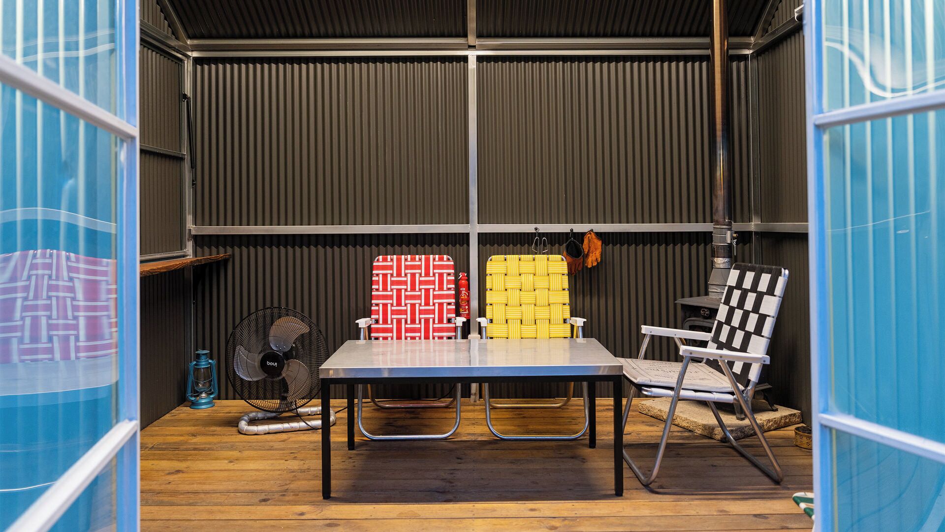 Habitación pequeña con una mesa, dos sillas, un ventilador y cajas de almacenamiento frente a una pared de metal corrugado.