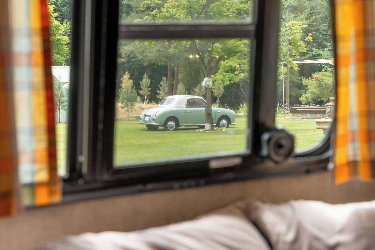 A window with a view of a green car parked on a grassy area, curtains, and a bed with a pillow.