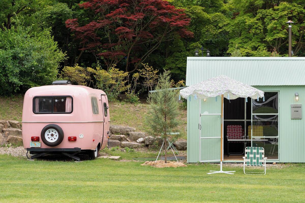 A pink RV parked in a grassy area with a small green shed and a white umbrella in front.