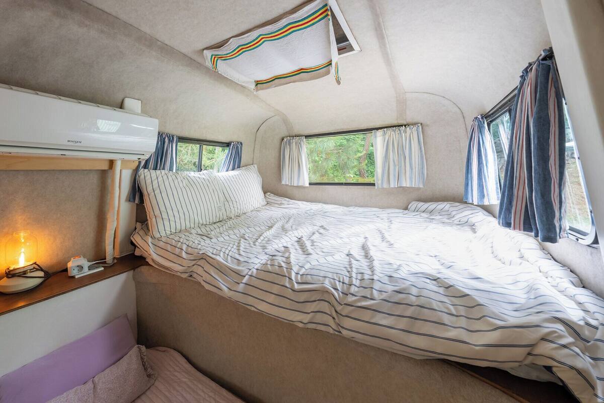 A cozy RV interior with a bed, a small table with a lamp, and a ceiling-mounted air conditioner.