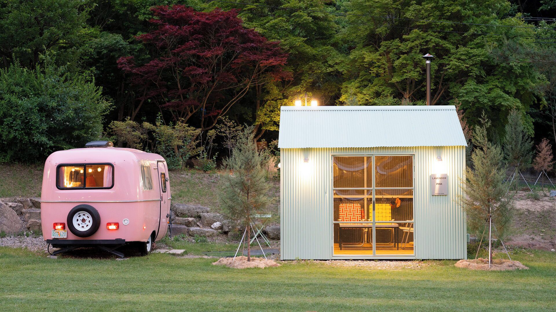 Una caravana rosa estacionada junto a un pequeño edificio de color claro con puerta de vidrio, rodeado de árboles.