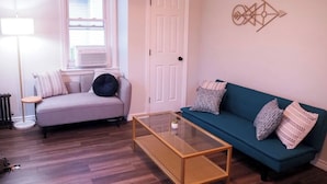 A living room with a grey sofa, a blue sofa, a glass coffee table, a white door, and a window with blinds. - Summer Home Close to Beach, Ocean, LIRR w/Balcony (Long Beach)