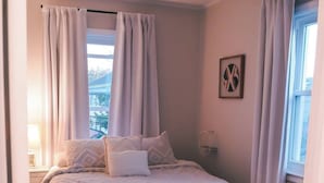 A bedroom with a bed, curtains, a bedside lamp, a wall clock, and a window with a view of buildings. - Summer Home Close to Beach, Ocean, LIRR w/Balcony (Long Beach)