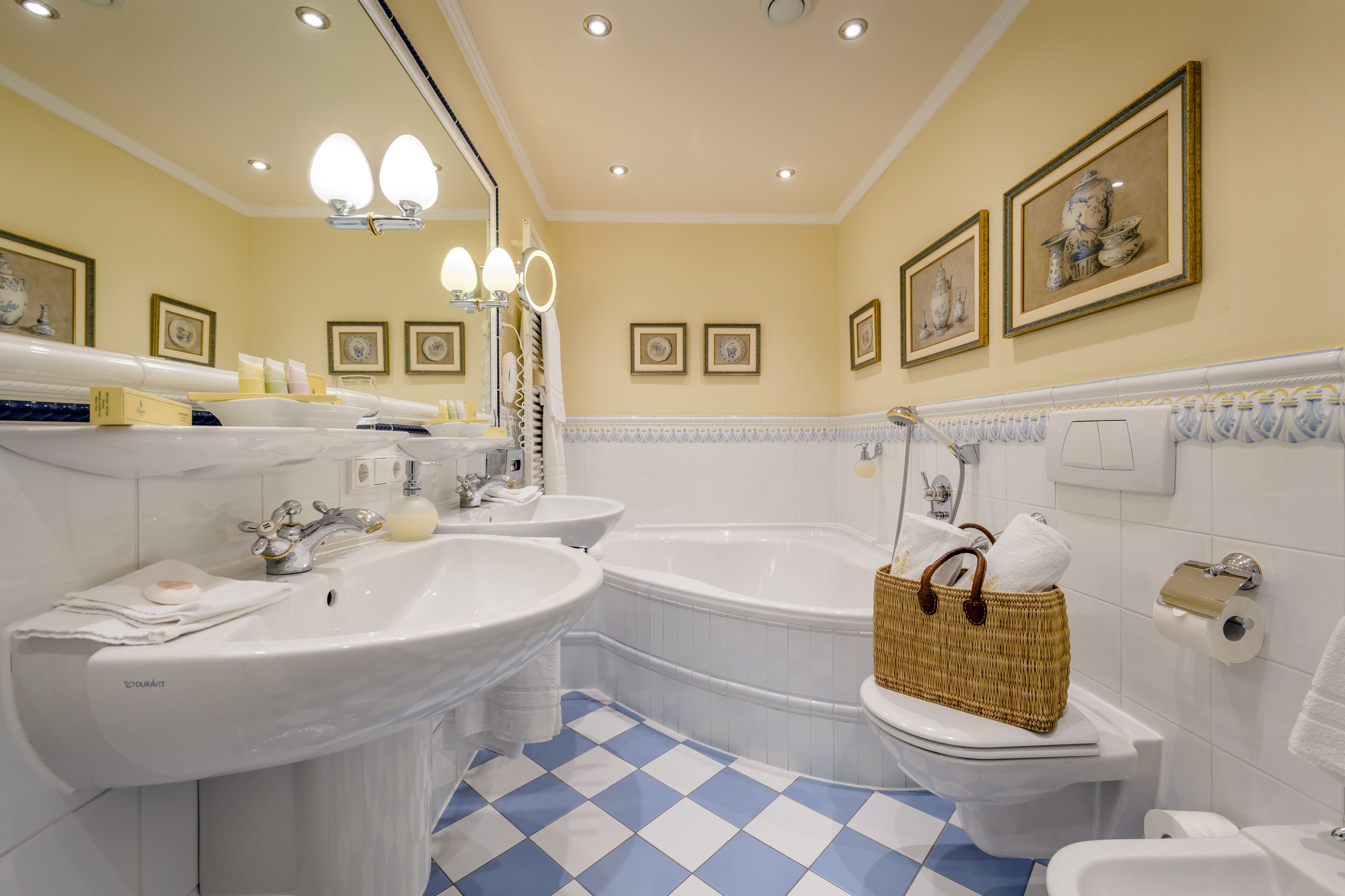 Une salle de bain avec deux lavabos, une baignoire et des toilettes. Des tableaux encadrés ornent le mur, un panier est posé sur les toilettes et le sol est recouvert de carreaux bleus et blancs.