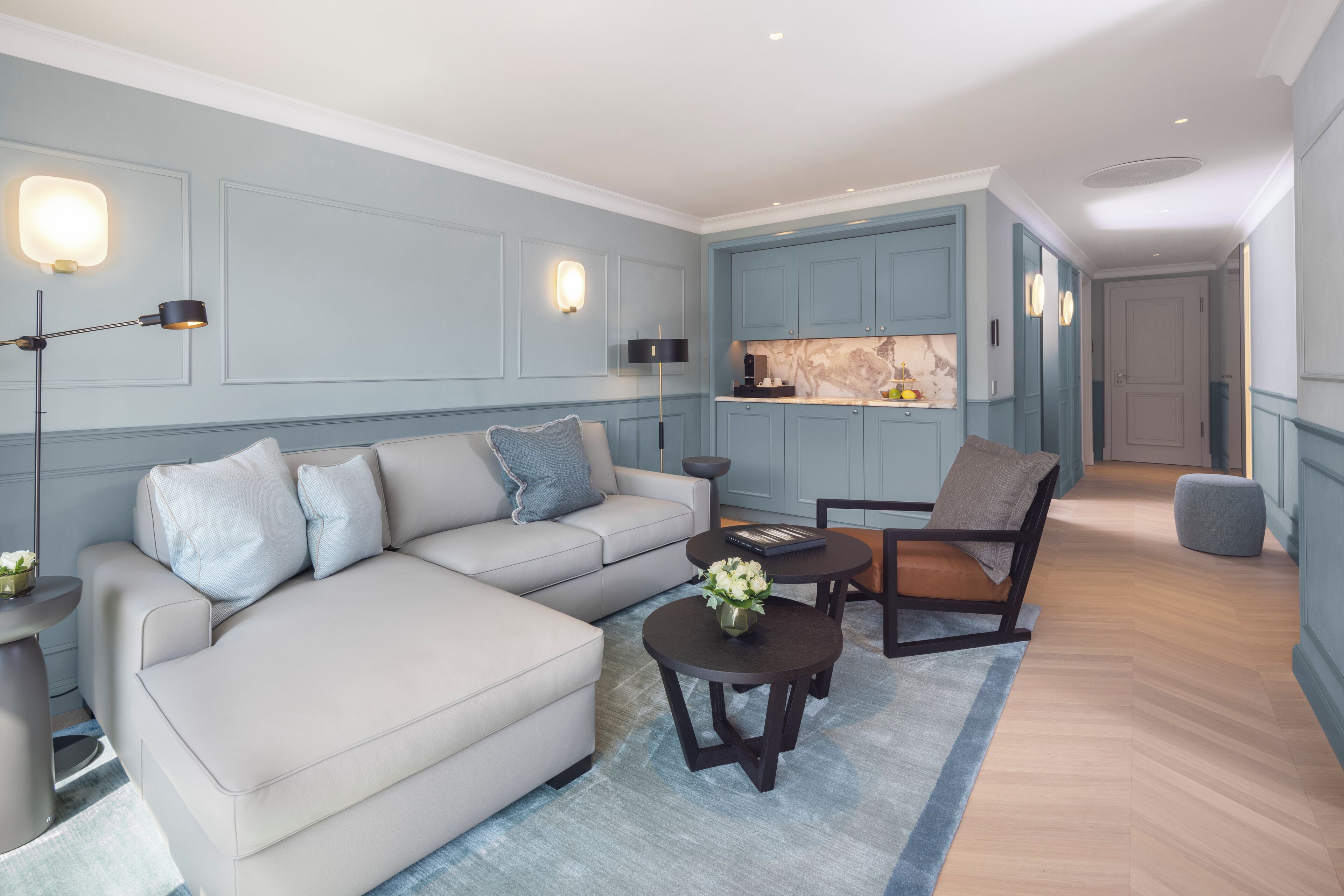 Una sala moderna con sofá, mesa de centro y sillón, un área de cocina con armarios azules, y un pasillo que conduce a una puerta.