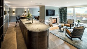 A modern kitchen with a central island, a dining area with chairs, a fireplace, and a large window with a view. - Hotel Bareiss (Baiersbronn)