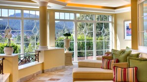 A spacious room with a large window offering a mountain view, a beige sofa with striped cushions, and a potted plant. - Hotel Bareiss (Baiersbronn)
