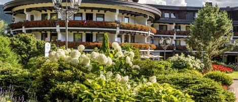 A hotel with a circular facade, multiple balconies with red flowers, and a well-maintained garden with various plants and white flowers.