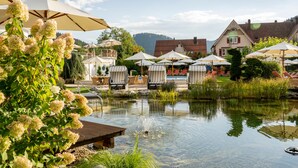 A lakeside setting with outdoor seating under umbrellas, a wooden deck, and a house in the background. - Hotel Bareiss (Baiersbronn)