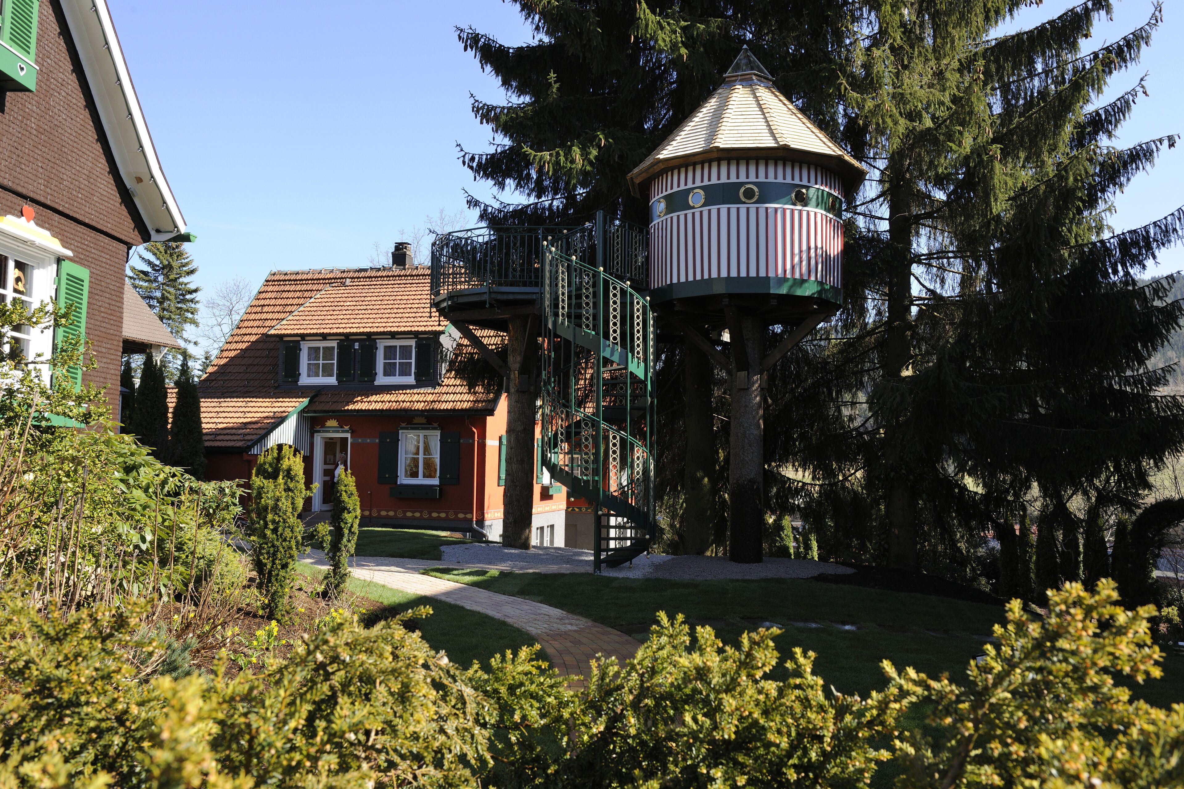 A traditional house with a red roof and green shutters, a small tower with a spiral staircase, and a well-maintained garden.